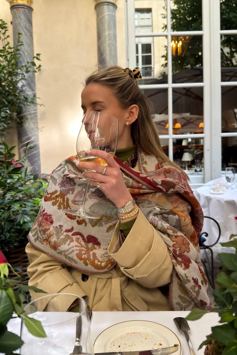 Annie in a camel coat and floral shawl with a glass of wine at a cafe table