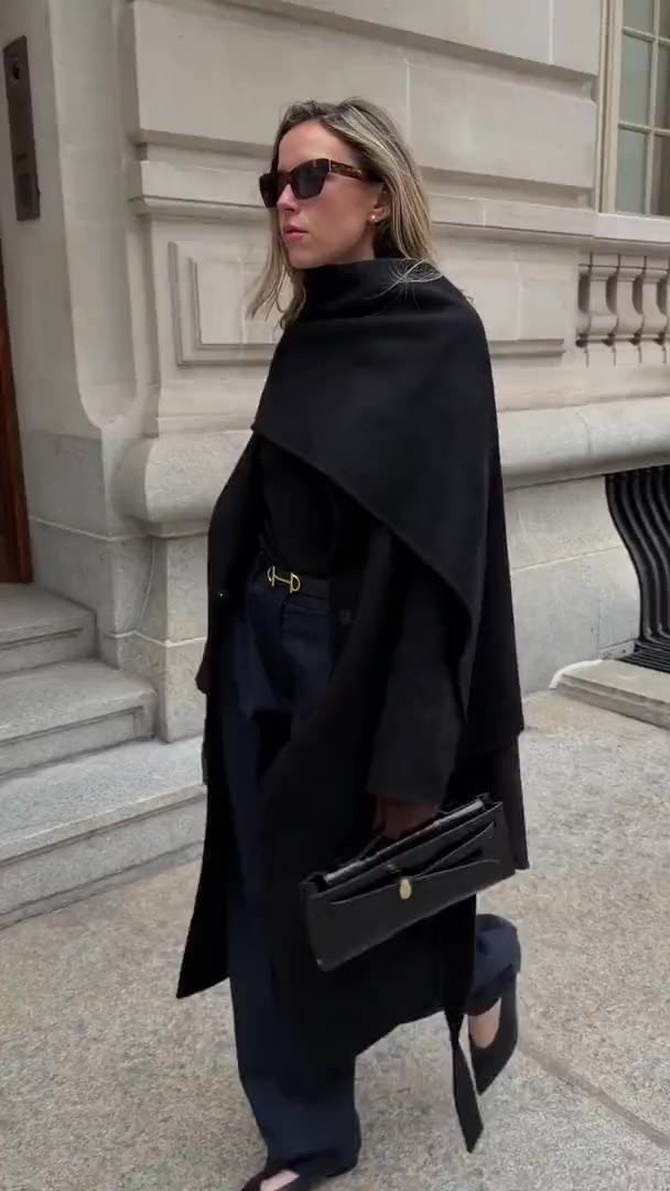 Annie walking past a limestone building in a black wrap coat, navy trousers and sunglasses